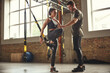 © Friends Stock - My personal trainer. Confident young man is showing slim athletic woman how to train legs with Trx fitness straps while training at gym.