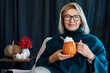 © okrasiuk - Smiling middle aged woman relaxing with pumpkin shaped cup of hot drink with whipped cream and cinnamon stick with fall mood composition for hygge home decor on background. Cozy autumn holidays.