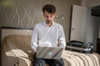 © Miljan Živković - One man adult caucasian businessman sit on the bed in hotel room work on his laptop computer prepare for meeting or remote online conference real person copy space wear white shirt
