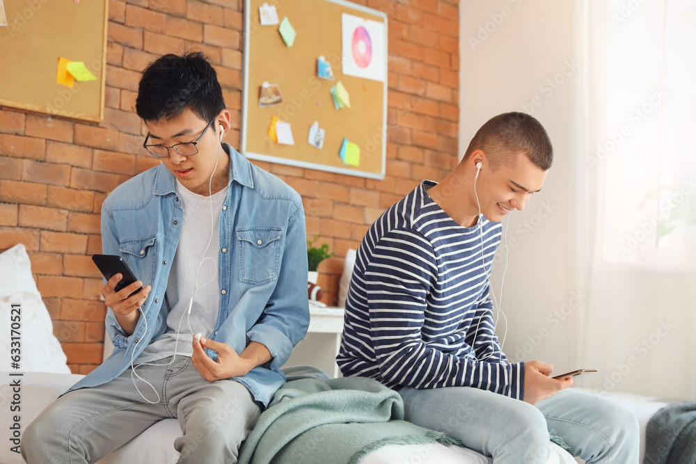 Male students with earphones using mobile phones in dorm room