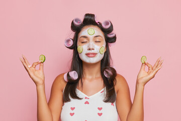 A brunette with curlers on her head and cosmetic mask on her face meditates with cucumbers in her hands, smiling calmly, face treatment concept, copy space