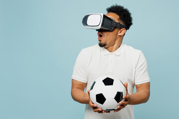 Wall Mural - Young amazed man fan wearing basic t-shirt cheer up support football sport team hold in hand soccer ball watching in vr headset pc gadget watch tv live stream isolated on plain blue color background.