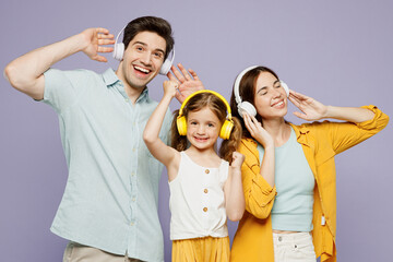 Wall Mural - Young happy smiling parents mom dad with child kid daughter girl 6 years old wear blue yellow casual clothes listen to music in headphones dance isolated on plain purple background Family day concept