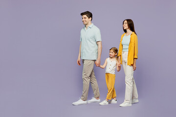 Full body side view young parents mom dad child kid daughter girl 6 years old wearing blue yellow casual clothes hold hands walk go look aside isolated on plain purple background. Family day concept.