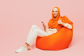 Wall Mural - Full body happy young arabian asian muslim woman wearing orange abaya hijab sit in bag chair hold use mobile cell phone isolated on plain pink background. Uae middle eastern islam religious concept.