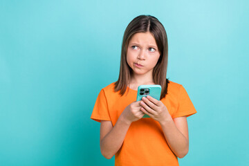 Portrait of puzzled confused schoolgirl dressed orange t-shirt look empty space smartphone in hands isolated on turquoise color background