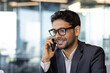 © Liubomir - Young successful arab businessman boss talking on phone close up, man smiling contentedly inside office at workplace in business suit