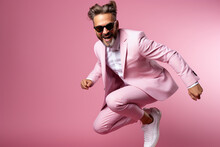 Young Handsome Funny Man With Glasses, Brown Hair And Beard, Wearing Light Grey Suit And Sneakers, Jumping With The Skateboard On Color Studio Background.
