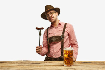 Bavarian mustache man in hat wearing traditional fest outfit holding hot fried sausage and looking at huge glass of beer. Oktoberfest concept