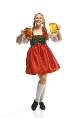 Wall Mural - Full-lenght portrait of friendly young woman wearing folk German dirndl with two beer mugs over white background. Holiday event, Oktoberfest