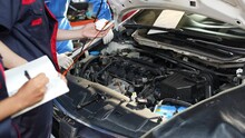 Selective Focus, Cropped View Of A Young Male Mechanic In Uniform, Standing In Front Of The Car With An Open Hood, Pulling Out The Dipstick And Wipe Off The Oil With A Cloth For Checking The Oil Level