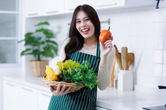 Happy portrait of young asian woman holding a basket of vegetables of standing a cheerful preparing food and enjoy cook cooking with vegetables, while standing on a kitchen Condo life or home