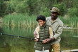 © AnnaStills - Happy African American dad catching fish together with his son on the lake outdoors