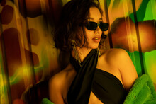 Pretty And Stylish Asian Woman In Sunglasses Standing Near Colorful Graffiti On Wall In Night Club