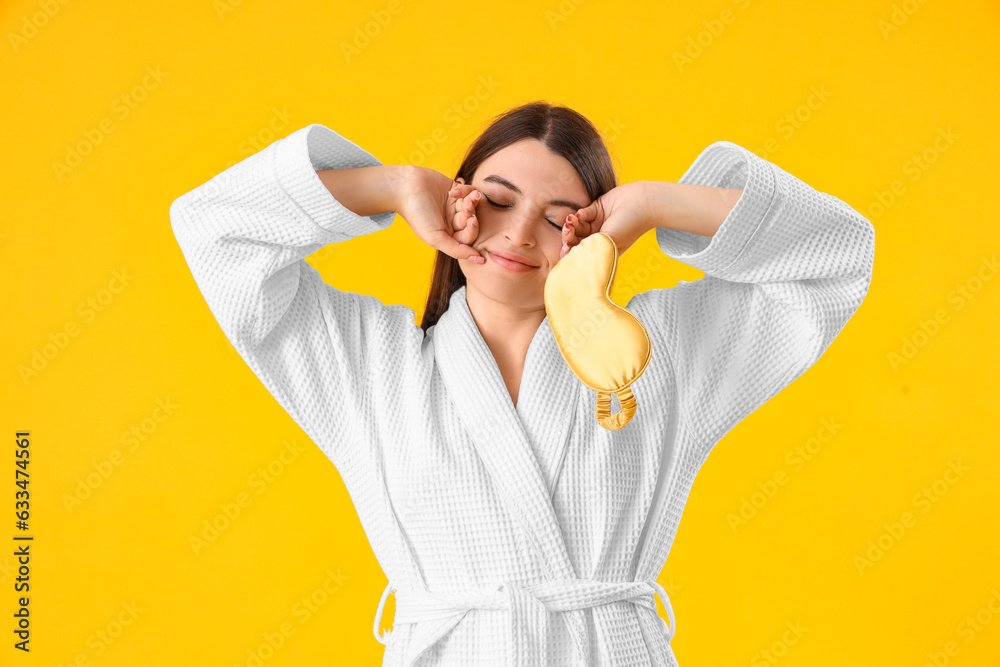 Young woman in gown with sleeping mask on yellow background