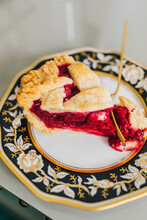 Single Slice Of Lattice-crust Raspberry Pie On White, Gold, Black Floral Plate With Gold Brass Fork Cutting Into Pie, Gray Table