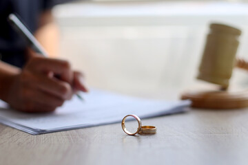 Wedding rings on wooden table with hand signing documents and judge gavel on the background. Marriage and divorce concept