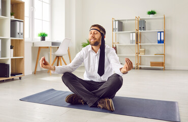 Funny, tired, stressed, young business man in a white shirt, with a necktie around his head sitting on a yoga mat on the office floor, doing meditation, and relaxing