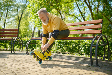 Wall Mural - Grey-haired senior man putting on roller skates while sitting on bench in park