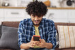 © kucherav - Happy young Asian man with curly hair using mobile phone at home, messaging or browsing social networks while relaxing sitting on sofa, smiling friendly