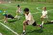 © pressmaster - African American schoolboy in activewear doing stretching exercise on green grass at stadium while his classmates repeating after him
