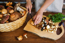 Women's Hands Cut Wild Mushrooms On A Wooden Board. There Is A Basket Of Mushrooms On The Table And Fresh Greens.