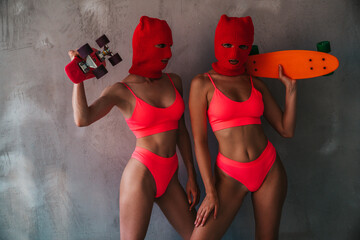 Two beautiful sexy women in red underwear. Models wearing bandit balaclava mask. Hot seductive female in nice lingerie near grey wall in studio. Crime and violence. Hold, penny skateboard