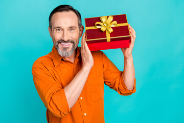 Sticker - Portrait of pleasant toothy beaming man dressed orange shirt hands hold present box near ear isolated on turquoise color background