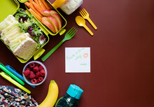 School Lunch Box With Sandwiches, Carrot Sticks, Apple, Banana, Lettuce, Hummus And Raspberries. Healthy School Lunch Concept. Burgundy Background And Note With Text - Have A Nice Day Mom. Top View 