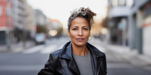 Headshot Portrait Of Stylish And Attractive Black Hispanic Queer Woman With Curly Short Hair And Gray Highlight Streaks, Wearing Black Leather Jacket, Standing In Urban Environment