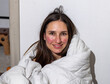 smiling woman with red spots on face, alcohol allergy hard light direct in face. girl wrapped in bed blanket sitting with hands on face. rosacea