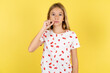 blonde kid girl wearing polka dot shirt over yellow studio background mouth and lips shut as zip with fingers. Secret and silent, taboo talking.