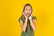 Stupefied blonde kid girl wearing green T-shirt over yellow studio background expresses excitement and thrill, keeps jaw dropped, hands on cheeks, has eyes popped out