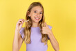 © Roquillo - blonde kid girl wearing violet T-shirt over yellow studio background holding an invisible braces aligner and rising thumb up, recommending this new treatment. Dental healthcare concept.