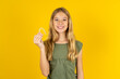 © Roquillo - blonde kid girl wearing green T-shirt over yellow studio background holding an invisible braces aligner, recommending this new treatment. Dental healthcare concept.