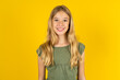 blonde kid girl wearing green T-shirt over yellow studio background with nice beaming smile pleased expression. Positive emotions concept