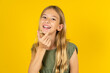 blonde kid girl wearing green T-shirt over yellow studio background holding an invisible aligner ready to use it. Dental healthcare and confidence concept.