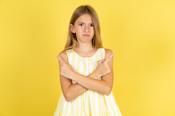 Serious young beautiful blonde kid girl over yellow studio background crosses hands and points at different sides hesitates between two items. Hard decision concept