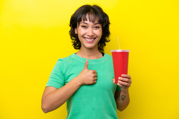 Wall Mural - Young Argentinian woman holding a soda isolated on yellow background giving a thumbs up gesture