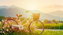 Bicycle With A Wicker Basket In A Beautiful Spring Landscape With Colorful Wildflowers In A Green Meadow, Mountain Background, At Sunset.Generative AI