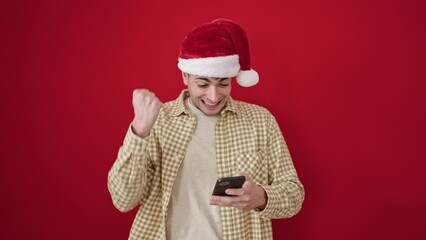 Sticker - Young hispanic man wearing christmas hat using smartphone celebrating over isolated red background