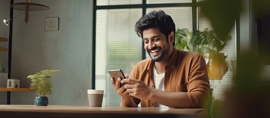 Happy Indian millennial browsing social networks on mobile phone and drinking coffee at home copy space