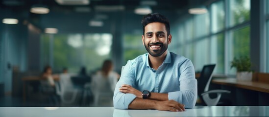 Sticker - Successful Indian male entrepreneur in a shirt smiling and looking at empty space sitting at his office desk with a panorama of the office interior and