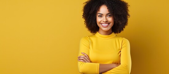 Young African American woman with confident and smiling demeanor posing on yellow background crossing arms on chest and maintaining eye contact with ca