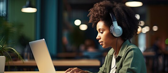 Black woman taking notes during office webinar using laptop and wearing headphones with empty space