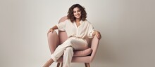 A Millennial Lady Posing And Relaxing Indoors Sitting In An Armchair Against A White Studio Wall Smiling At The Camera In A Full Length Portrait