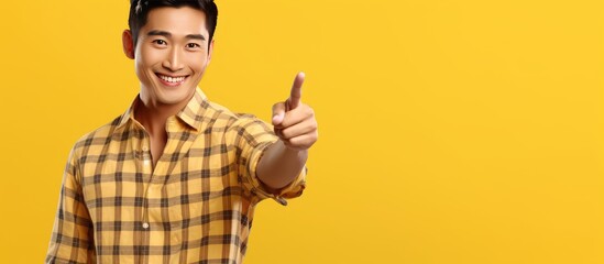 Poster - Young attractive Asian man in his 20s smiling wearing a plaid shirt pointing at an advertisement on a yellow background representing a lifestyle concep