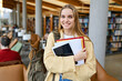 © insta_photos - Smiling cute pretty blond girl, positive female teenage high school student holding backpack looking at camera standing in modern university or college campus library, portrait.