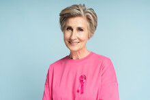 Portrait Of Confident Smiling Senior Woman Wearing Pink T Shirt With Pink Ribbon Isolated On Blue Background. Health Care, Support, Prevention. Breast Cancer Awareness Month Concept