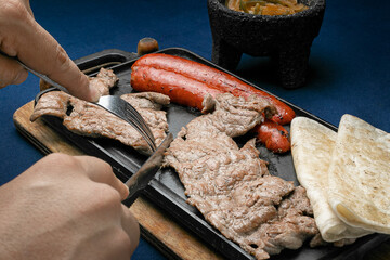 dish of Mexican roast beef and pork, accompanied by two quesadillas and molcajete sauce, on a wooden board and a matal comal on a blue table.